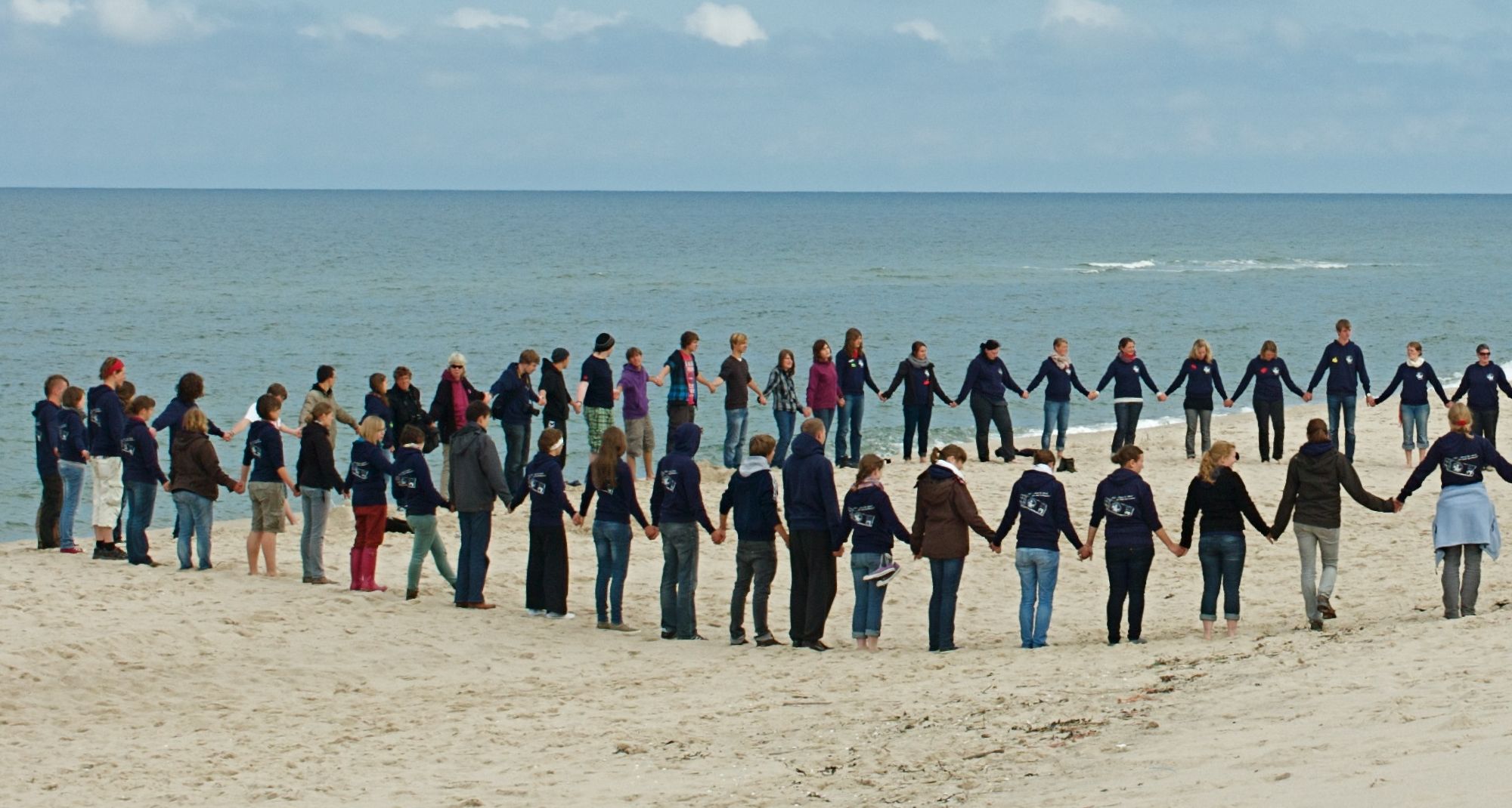 Eine große Gruppe an Menschen steht Hände haltend im Kreis am Strand